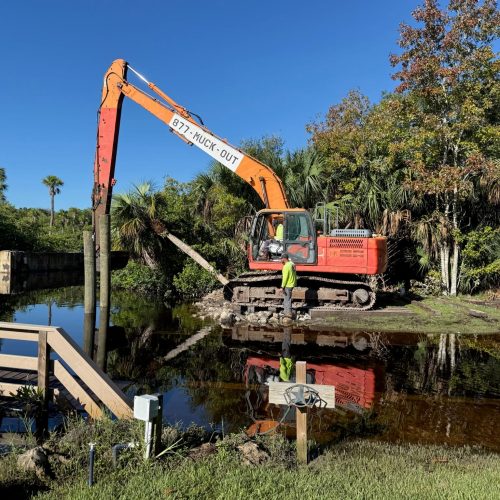 A serene waterfront scene with a dredging vessel in the foreground, calm water reflecting the clear blue sky.